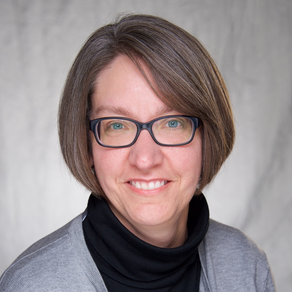 Jenniffer Carpe, a woman with short hair and glasses, wearing a black turtleneck and grey sweater.