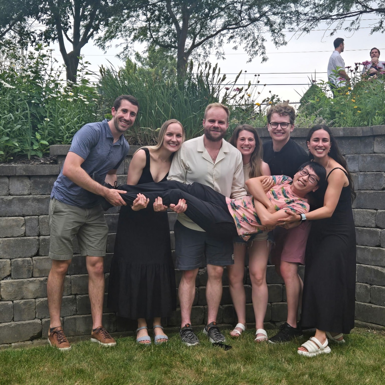Standing outside by a stone wall, five residents stand together and hold Dr. Cheung, who is laying across their arms.